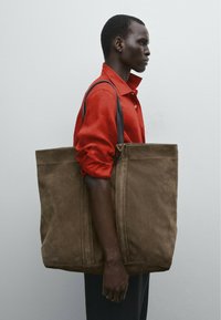 Sac en daim marron avec deux poignées, forme carrée et lignes épurées. Modèle portant une chemise rouge se tient devant un fond uni.