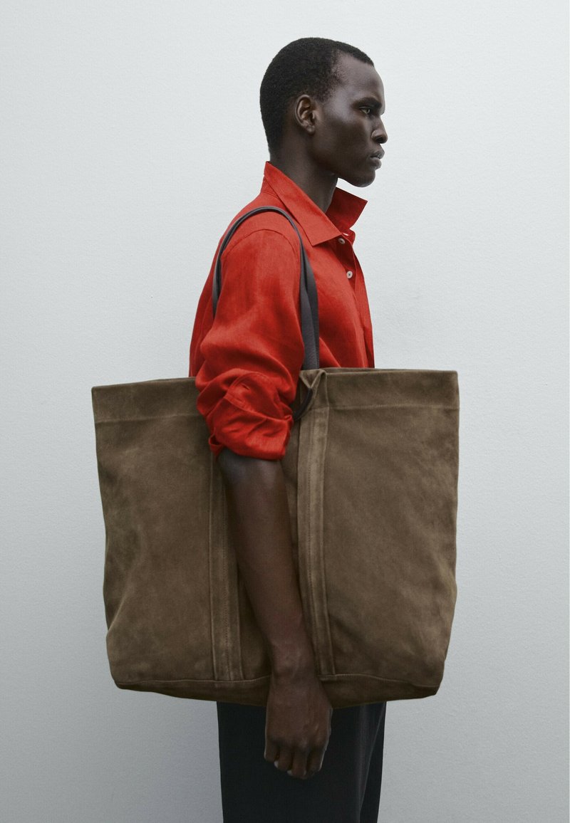 Sac en daim marron avec deux poignées, forme carrée et lignes épurées. Modèle portant une chemise rouge se tient devant un fond uni.