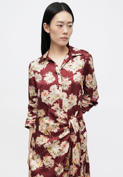 Czerwona sukienka w kwiaty z satyny o gładkiej fakturze. Posiada kołnierzyk, zapięcie na guziki z przodu oraz wiązanie w talii, a jej wzór to delikatne pastele.