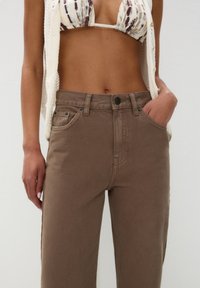 Pantalon en denim marron à taille haute, doté de poches avant, de surpiqûres ton sur ton et d'une fermeture à bouton. Texture lisse, design décontracté.