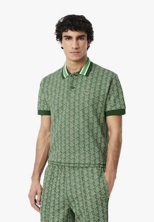 Grönmönstrad polotröja med geometrisk design, med kontrasterande vit och grön randig krage och manschetter. Lacoste-logotyp på bröstet.