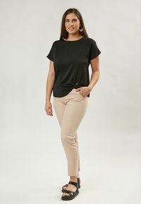 Top nero a maniche corte con dettaglio annodato, abbinato a pantaloni attillati beige chiaro. La modella indossa sandali neri; sfondo neutro.