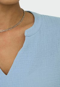 Lyseblå, struktureret stof med V-udskæring og en blød, let krøllet overflade. Har en rund krave og et sølvfarvet kædenecklace.