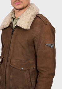 Giacca di pelle marrone con colletto in shearling beige, chiusura con zip, tasche laterali e una patch decorativa sulla manica sinistra.