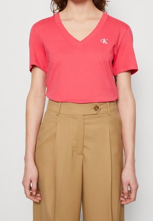 Femme portant un t-shirt corail à manches courtes et col en V avec le logo "cK" et un pantalon taille haute couleur camel avec une ceinture boutonnée.