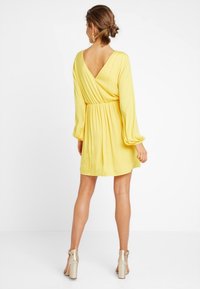 Vestido envolvente amarillo con mangas largas abullonadas, espalda en forma de V y cintura elástica, combinado con sandalias metálicas de tacón bloque.