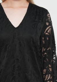 Μαύρη δα�ντελένια μπλούζα με V-λαιμό και μακριά μανίκια, με περίπλοκα λουλουδάτα σχέδια και υφή υφάσματος. Διαφανείς λεπτομέρειες στα μανίκια.