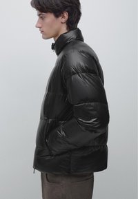 Młody mężczyzna z profilu, ubrany w czarną błyszczącą kurtkę puchową, ręce w kieszeniach, na jednolitym jasnym tle.