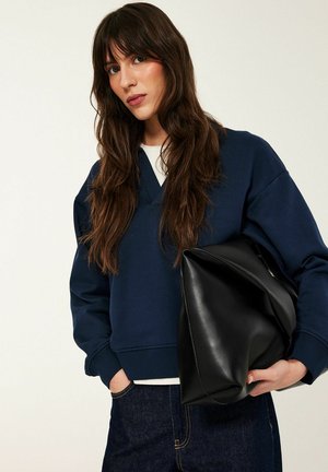 Frau mit langem braunem Haar trägt ein marineblaues Sweatshirt und Jeans, hält eine große schwarze Leder-Clutch vor einem einfarbigen Hintergrund.