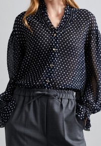 Blouse bleu marine transparent à pois blancs, col à volants et manches bouffantes. Boutons dorés agrémentent le devant. Associée à un pantalon gris.