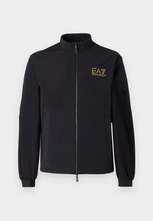 Giacca softshell nera con colletto alto, chiusura con zip frontale e due tasche laterali. Presenta un logo dorato EA7 Emporio Armani sul petto.