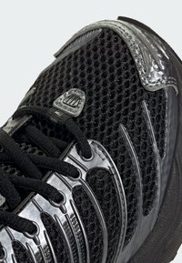 Čierna športová obuv s textúrou zo sieťoviny, striebornými akcentmi a zakriveným dizajnom. Obsahuje zložité detaily a čierne šnúrky. Štýl zameraný na pohodlie.