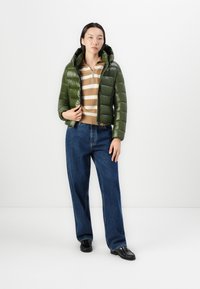 Groene gewatteerde jas met capuchon, gestreepte bruin en crèmekleurige trui eronder, brede blauwe spijkerbroek en zwarte loafers. Eenvoudig, functioneel ontwerp.