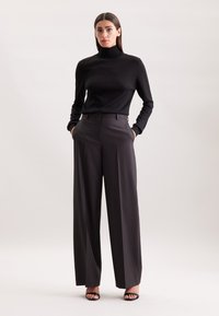 Donna in piedi con le mani in tasca, che indossa un dolcevita nero e pantaloni larghi grigio scuro, con tacchi aperti neri.