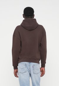 Καφέ φούτερ με κουκούλα και άνετη εφαρμογή, με ριμπ μανσέτες, φορεμένο με ανοιχτό μπλε τζιν με εφέ φθοράς. Λείος υφασμάτινος, με ελάχιστες λεπτομέρειες.