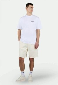 Λευκό βαμβακερό T-shirt με κοντά μανίκια και ένα μικρό μαύρο λογότυπο, συνδυασμένο με μπεζ σορτς. Λείου υφής, με casual εφαρμογή και ουδέτερα χρώματα.