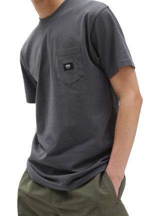 Mężczyzna w ciemnoszarym t-shircie Vans z przednią kieszenią i oliwkowozielonych spodniach, ręce w kieszeniach, na białym tle.