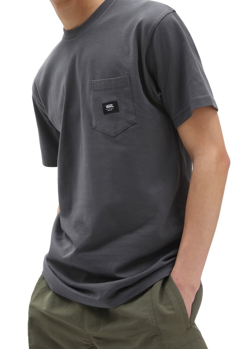 Homme portant un t-shirt Vans gris foncé avec poche avant et un pantalon vert olive, mains dans les poches, sur fond blanc.