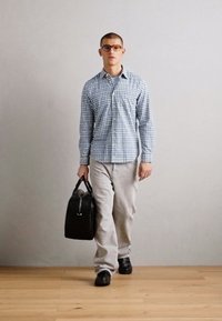 Černá kožená taška, modro-bílá kostkovaná košile, světle šedé kalhoty, elegantní černé boty. Neutrální pozadí a dřevěná podlaha.