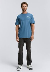 Sininen lyhythihainen t-paita pyöreällä kaula-aukolla ja valkoisella logotekstillä rinnassa; yhdistetty tummiin farkkuihin ja monivärisiin lenkkareihin.