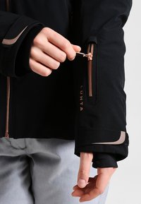 Schwarze Jacke mit tailliertem Design, ausgestattet mit einer Reißverschlusstasche und kontrastierenden roségoldenen Beschlägen. Strukturierten Bündchen sorgen für zusätzlichen Komfort.
