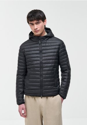 Jonge man die een zwart gewatteerd jack met capuchon en beige broek draagt, staand met één hand in zijn zak tegen een effen achtergrond.