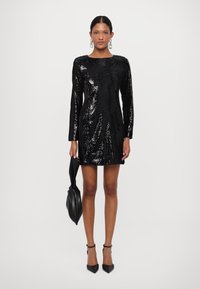 Mini abito nero con paillettes, maniche lunghe e scollo rotondo. La modella tiene una borsa nera, indossando tacchi a punta neri. Tocco morbido.