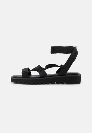Sort flad sandal med tykke justerbare remme over tæerne, midtfoden og omkring anklen, med rillede såler.