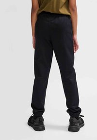 Schwarze Jogginghose mit elastischem Bund und ankleinen Bündchen, kombiniert mit schwarzen Sportschuhen. Der Stoff wirkt glatt und flexibel.