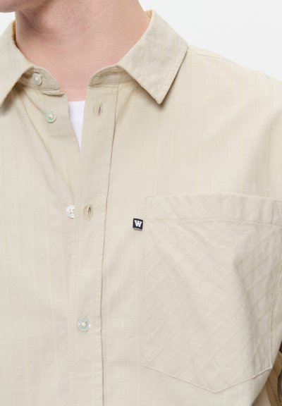Chemise boutonnée beige à rayures avec col, poche poitrine simple et un petit patch logo noir. Tissu doux avec une texture subtile.