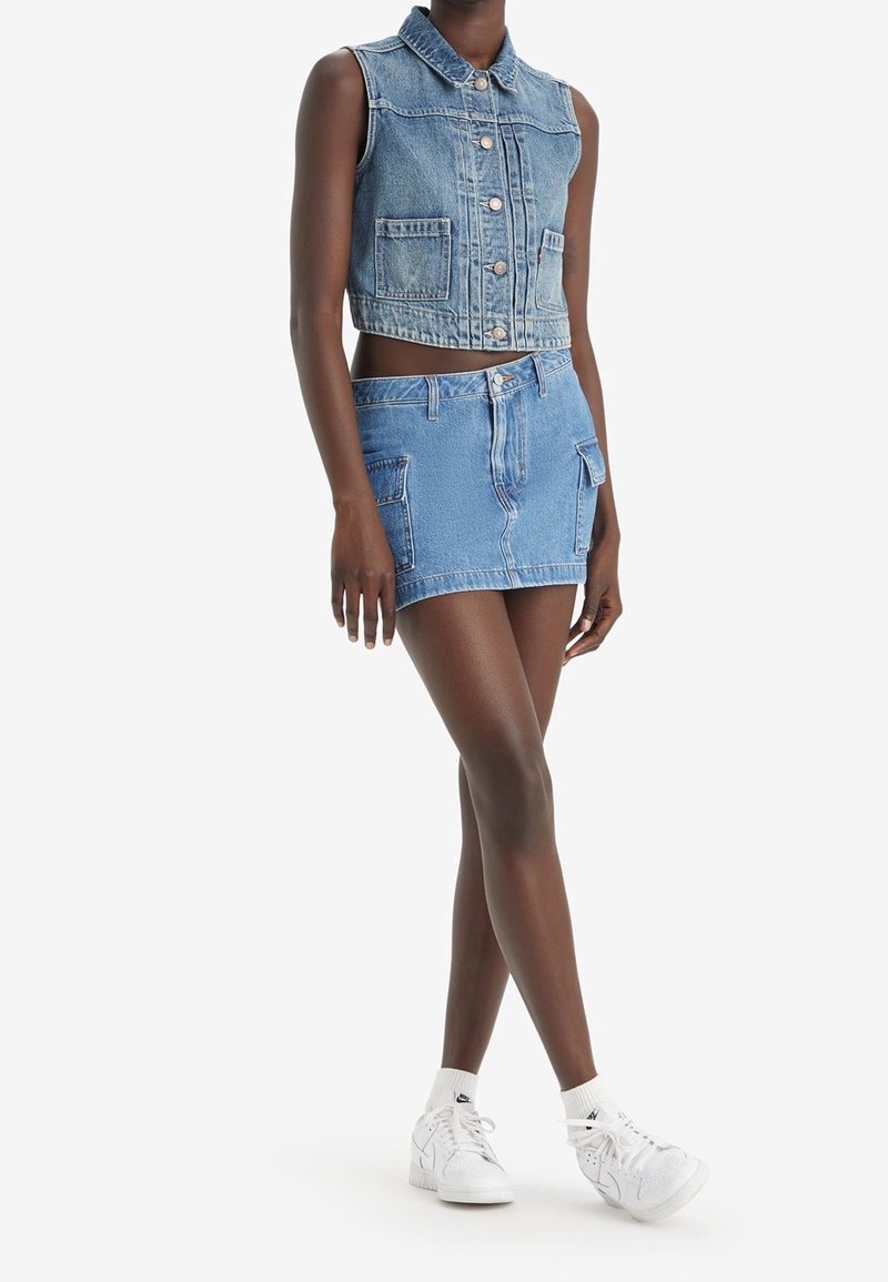 Ljusblå denimcropväst med ärmlös design, framknappar och två bröstfickor; i kombination med en matchande denimminikjol med sidfickor.