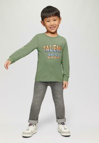 Grünes Langarmshirt mit dem Aufdruck "TALENT 2024" in mehrfarbigem Text, kombiniert mit grauen Jeans und weißen Sneakers mit grauen Akzenten.