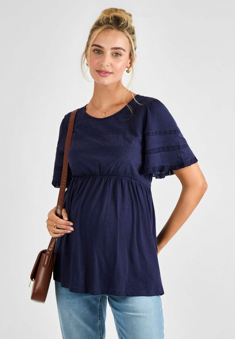 Tengerészkék, rövid ujjú felső, gyűrt derékrésszel, csipke díszítéssel és kerek nyakkivágással. Világoskék farmernadrággal és barna keresztpántos táskával párosítva.