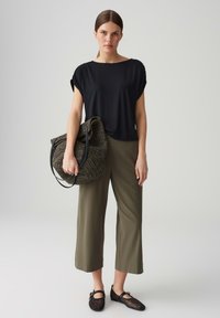 Schwarzes Kurzarmoberteil, olivfarbene Cropped-Hose, geflochtene dunkle Tasche mit schwarzen Griffen und schwarze Netzsandalen, vor einem neutralen Hintergrund stehend.
