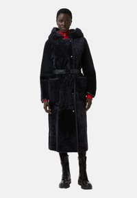 Mørkeblå faux fur frakke med hætte, to frontlommer, taljebånd og sølvfarvede knaplukninger. Båret over en rød zip-up trøje.