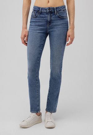 Blaue Denim-Jeans mit schmaler Passform, mittelhohem Bund, fünf Taschen und dezenten Nähdetails. Kombiniert mit weißen Sneakers.