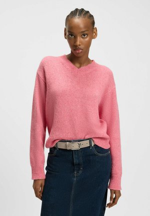 Maglione rosa a v scollo realizzato in un tessuto morbido e texturizzato, abbinato a una gonna in denim scuro e una cintura di colore chiaro. Design semplice con maniche lunghe.