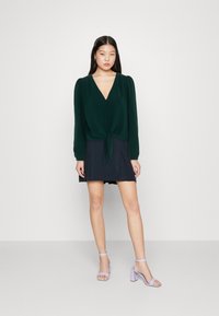 Green blouse with long sleeves, V-neck, buttoned front, and tied hem. Paired with a dark skirt and light purple block-heel sandals.