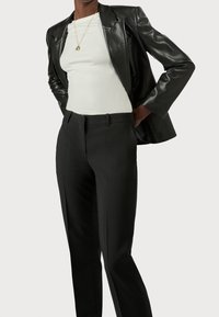 Person wearing black leather blazer over white top and black tailored pants, hands resting on lower back against a plain background.