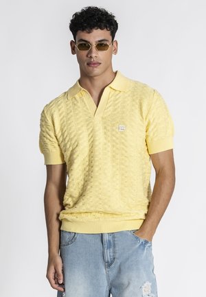 Mand iført sekskantede solbriller, tekstureret gul kortærmet polo, og lyseblå revnede cowboyshorts, stående med den ene hånd i lommen.