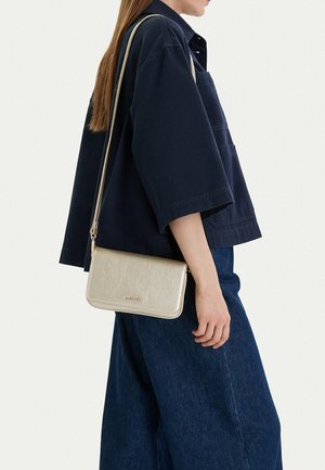 Osoba nosící tmavě modrou volnou košili a široké džíny, přehodila si přes rameno malou zlatou kovovou kabelku přes tělo.