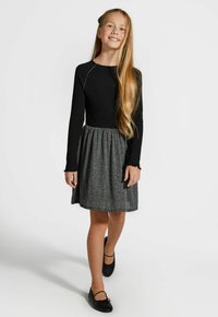 Czarna, długorękawowa bluzka z prążkowanej tkaniny zestawiona z szarym, teksturowanym spódnicą. Czarne płaskie buty dopełniają strój. Gładkie włosy spięte czarną spinką.