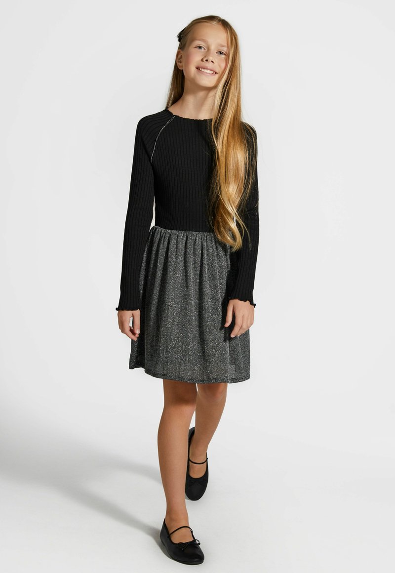 Czarna, długorękawowa bluzka z prążkowanej tkaniny zestawiona z szarym, teksturowanym spódnicą. Czarne płaskie buty dopełniają strój. Gładkie włosy spięte czarną spinką.