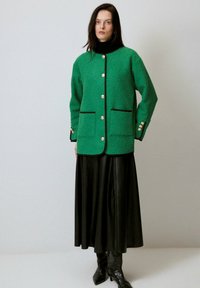 Jachetă texturată verde cu nasturi aurii, margine neagră și două buzunare frontale, asociată cu o fustă lungă din piele neagră și cizme până la genunchi.