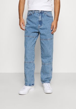 Mężczyzna ubrany w jasnoniebieskie jeansy z poziomymi przeszyciami i białe trampki, stojący na tle jednolitego białego tła.