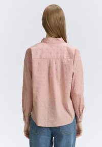 Camicia rosa testurizzata con motivo a pois in rilievo, maniche lunghe, polsini abbottonati e un bavero posteriore, abbinata a jeans azzurri chiari.