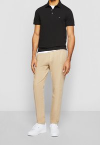 Man i svart kortärmad polo, beige byxor och vita sneakers som står mot en enhetlig ljus bakgrund.