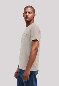 Beige cotton t-shirt featuring green graphics and text, short sleeves, a crew neckline, and a loose fit, worn with blue denim jeans.