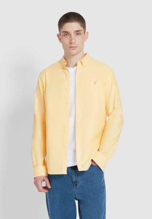 Lichtgele button-up shirt met een kraag, lange mouwen en een klein geborduurd logo, gedragen over een wit T-shirt en blauwe spijkerbroek.