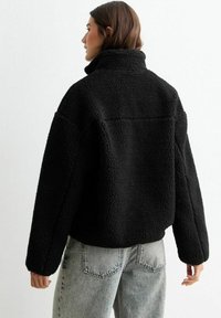 Svart fleecejacka med hög krage och texturerad yta. Lätt lös passform och cropped design, kombinerad med ljusgrå denimjeans.